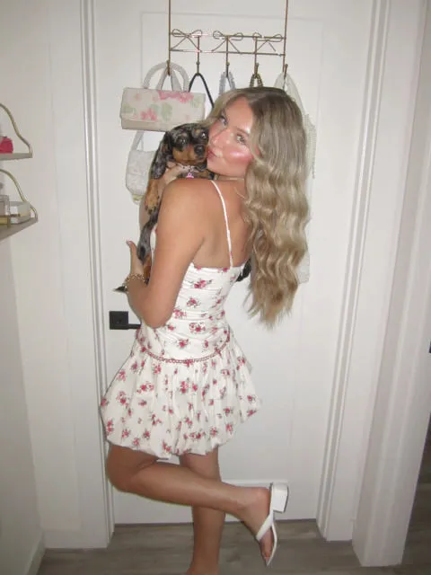 Macie in a white floral-print mini dress with a corseted bodice, holding her dachshund beside a wall of hanging handbags.