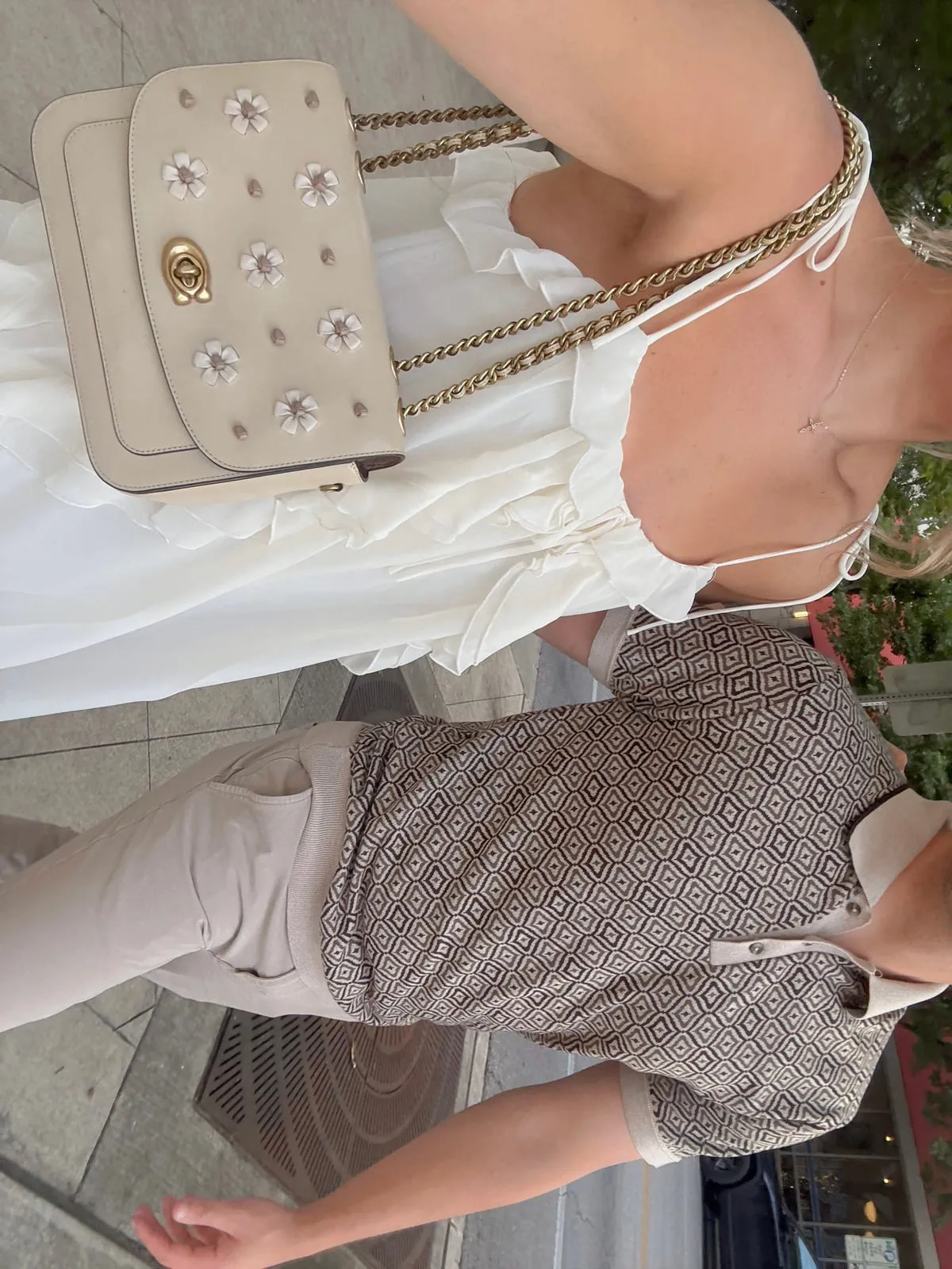 Overhead angle of Macie in a white ruffled dress, carrying a cream Coach bag with floral appliqué on a gold chain strap.