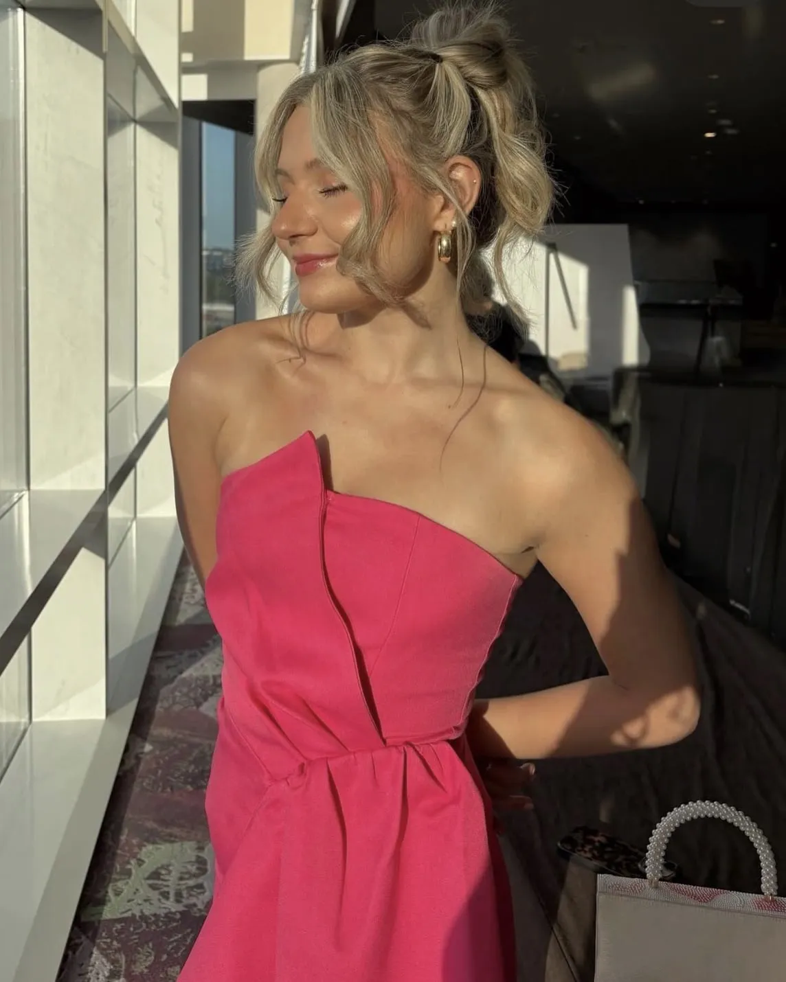 Macie in a hot pink strapless dress with a gathered waist, gold hoop earrings and a loose updo, turning into golden-hour light beside a window wall.