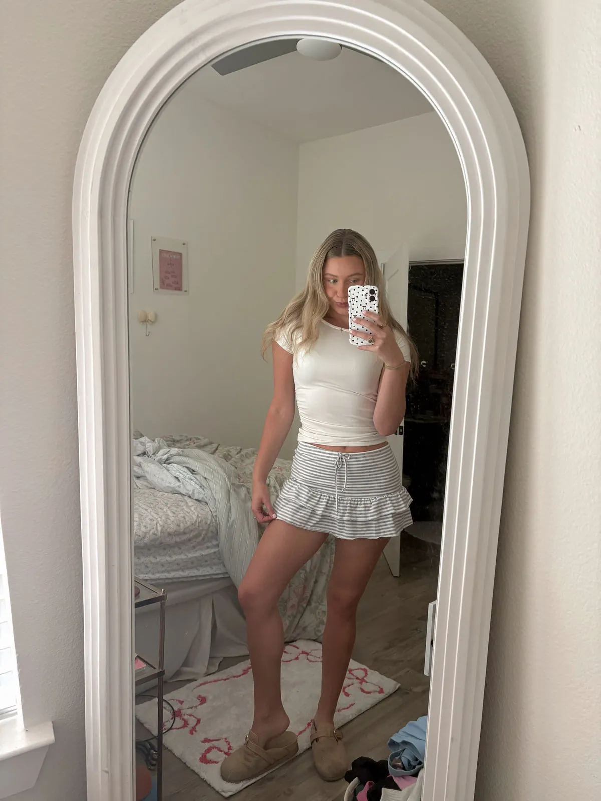 Macie in a fitted white tee with a white tiered ruffle mini skirt and woven flats, reflected in a tall arched mirror.