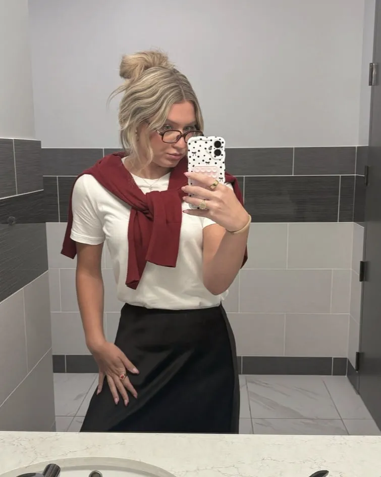 Macie in a white tee with a dark red sweater tied over the shoulders and a black satin midi skirt, wearing glasses.
