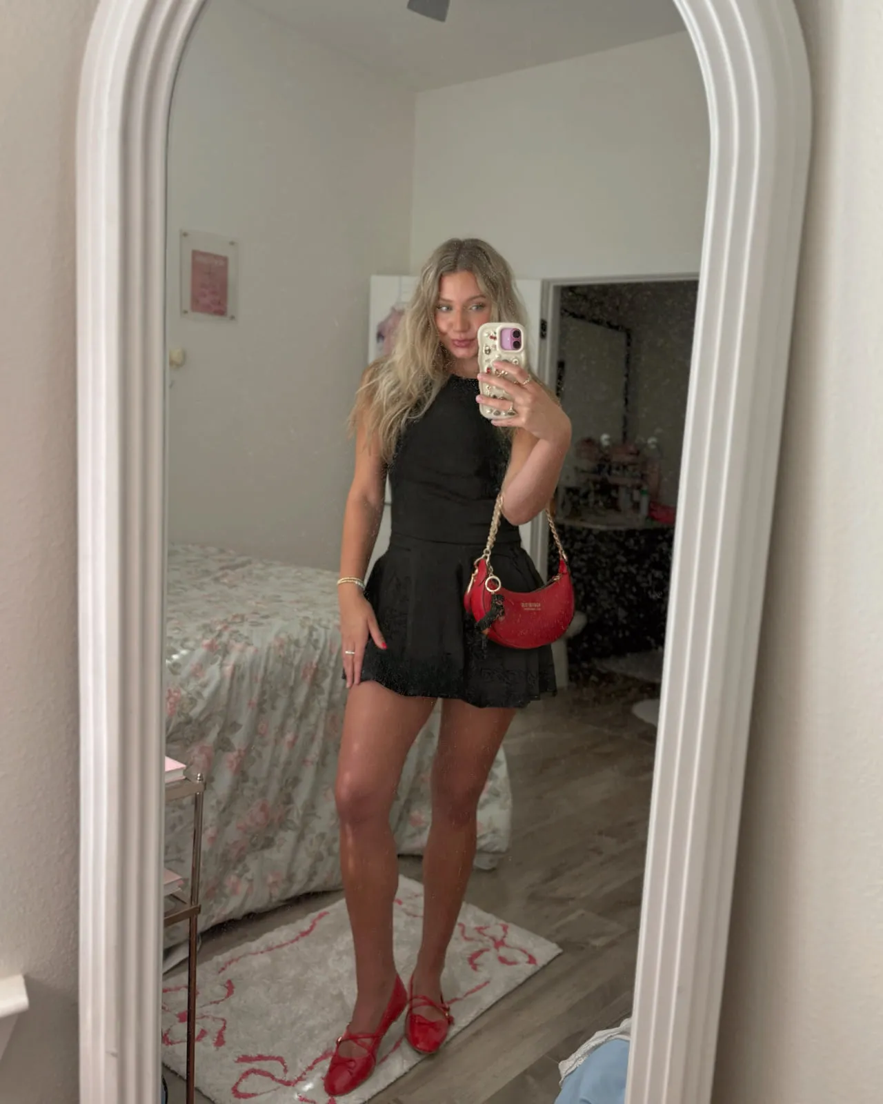 Macie in a black sleeveless romper with a red shoulder bag and red mary jane flats, reflected in a tall arched mirror.