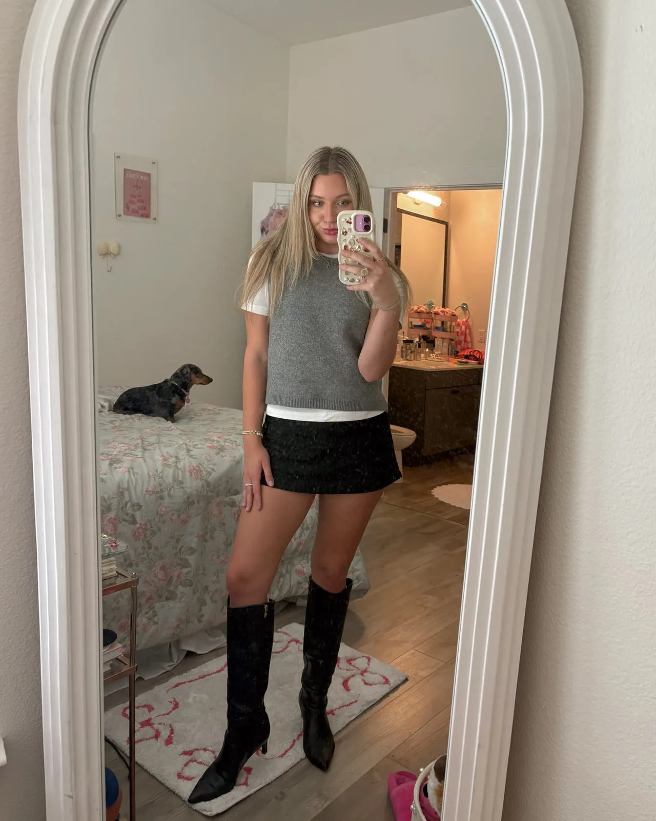 Macie in a grey sweater vest over a white tee with a black mini skirt and black knee-high boots, reflected in a tall arched mirror.