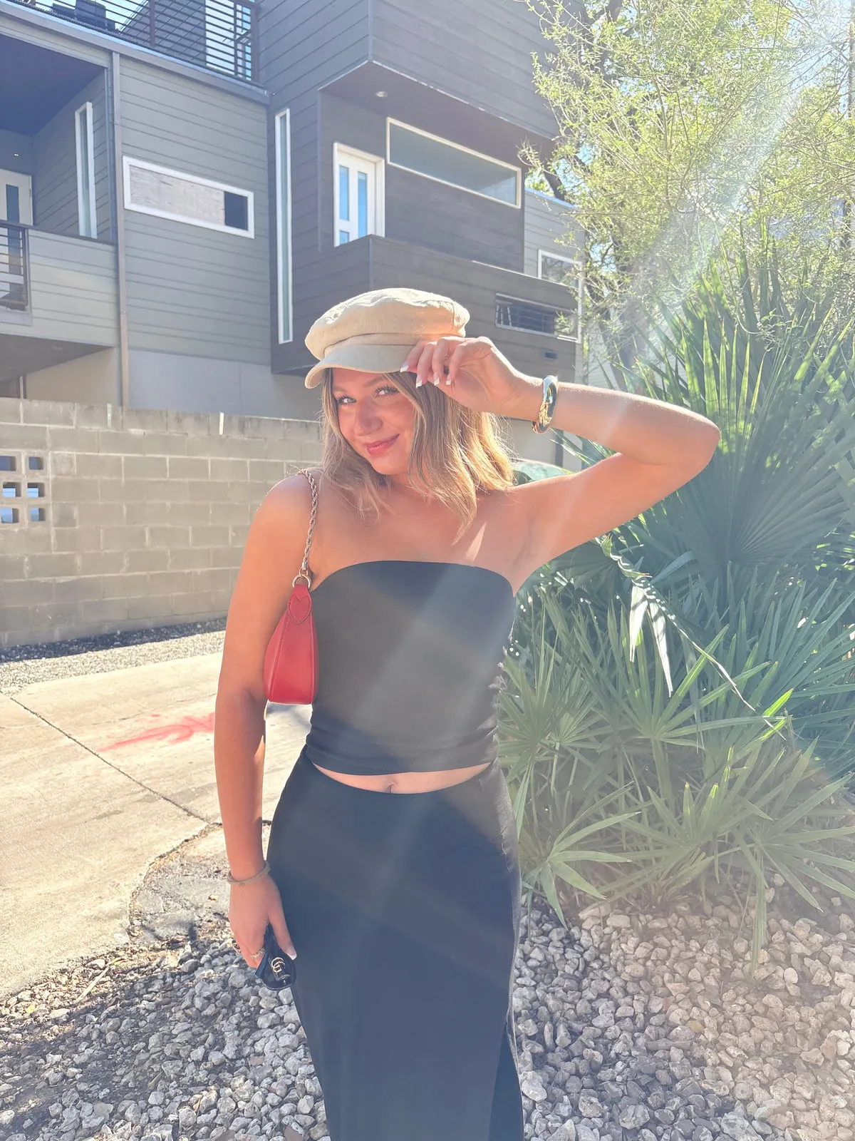 Macie in a black strapless column top and matching floor-length skirt with a tan newsboy cap and a red shoulder bag, backlit by afternoon sun outside a modern building.