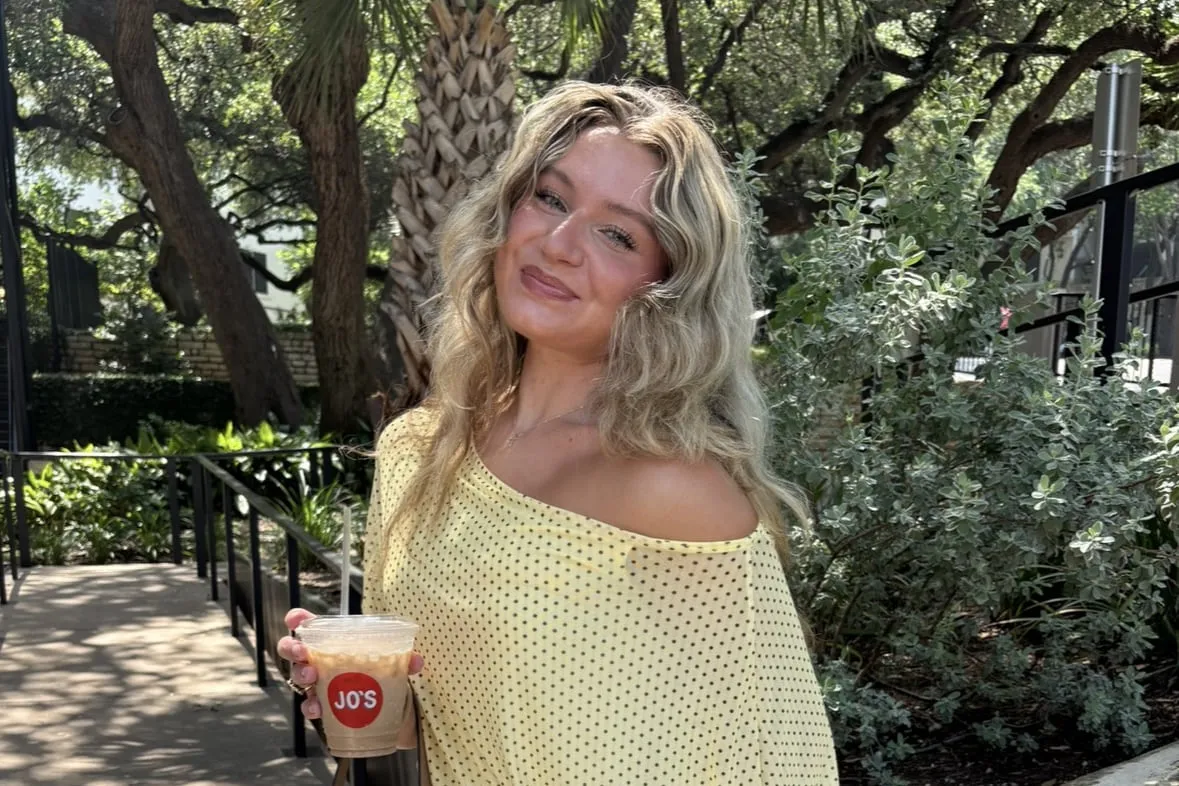 Macie in a pale yellow off-shoulder polka-dot top, holding an iced coffee on a shaded garden path.