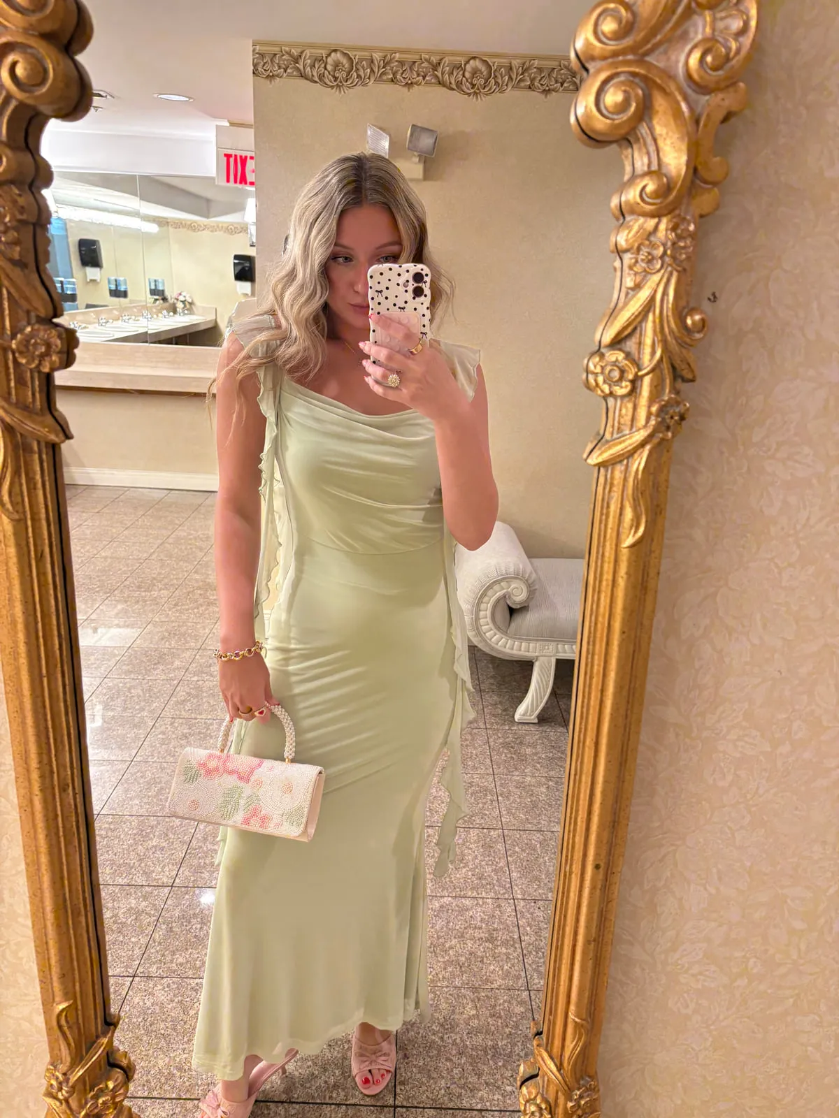 Macie in a sage green satin slip dress with a floral-print top-handle bag, standing in front of an ornate gold mirror.